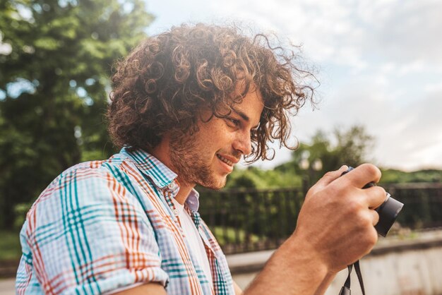 写真 彼のデジタルカメラで写真をチェックする巻き毛を持つ白人男性の肖像画都市公園に立っているデジタル写真カメラでカジュアルなシャツを着ている若いハンサムな男性人々の旅行とライフスタイル