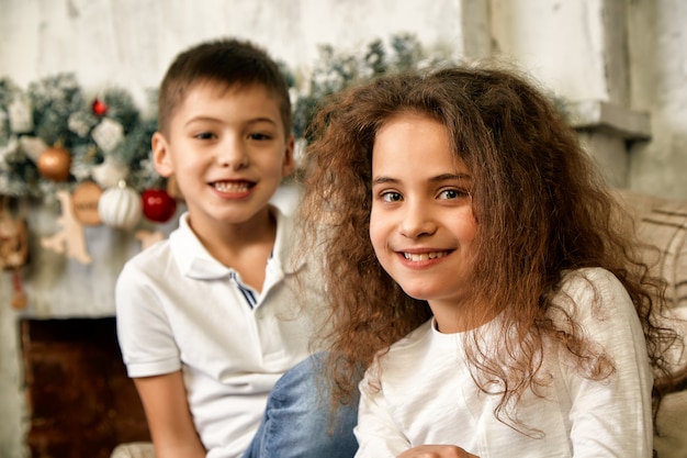 写真 新年の装飾で飾られた暖炉のそばの贈り物を待っているクリスマスの子供たちの肖像画。コンセプトクリスマスと奇跡を待っている子供たち。