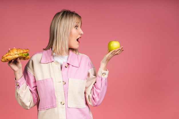 Фото Портрет смущенной молодой блондинки, выбирающей между круассаном и зеленым яблоком, изолированным на розовом фоне. концепция нездоровой пищи и диеты.
