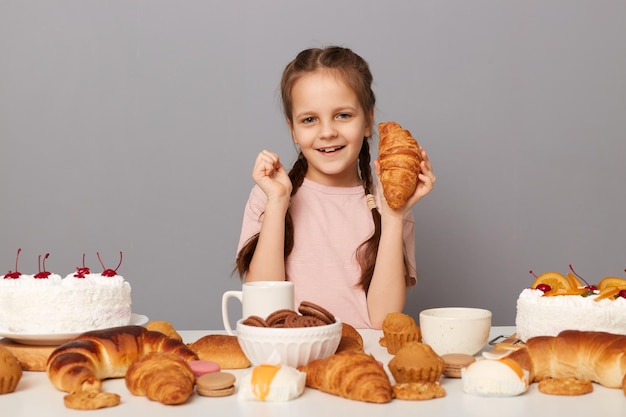 Портрет милой очаровательной счастливой девочки с косичками, сидящей за столом со сладкими десертами, изолированными на сером фоне, держащей круассан в руке и смотрящей в камеру