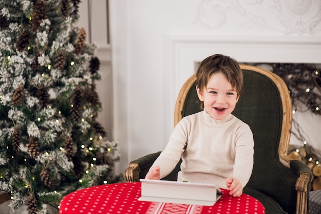 Портрет милого мальчика ребенк сидя на зеленом стуле и играя с таблеткой компьютера во время времени рождества. праздники сезонов.