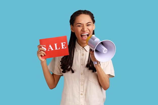 Фото Портрет эмоциональной женщины с черными дредами, держащей карточку с надписью о продаже и кричащей в мегафон о скидках, в белой рубашке. крытая студия снята на синем фоне.