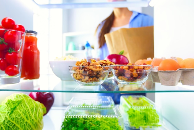 Фото Портрет женщины, стоящей возле открытого холодильника, полного здоровой пищи, овощей и фруктов
