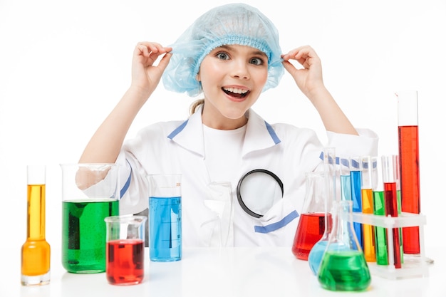 Фото Портрет забавной маленькой девочки в белом лабораторном халате, проводящей химические эксперименты с разноцветной жидкостью в пробирках, изолированных на белой стене