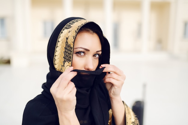사진 야외에서 서 있고 카메라를 보면서 스카프로 그녀의 얼굴을 숨기고 화려한 이슬람 여자의 초상화.