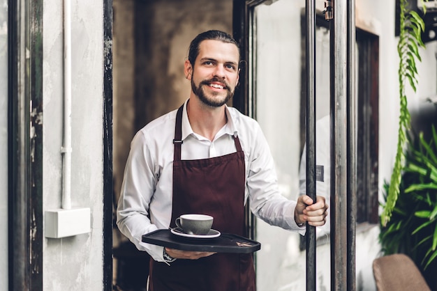 Фото Портрет красивого бородатого бариста человек мелкого предпринимателя, улыбаясь и держа чашку кофе в кафе или кафе. мужской бариста стоит в кафе
