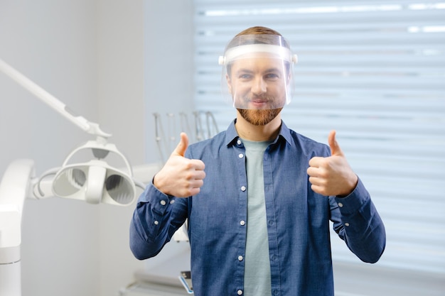 Фото Портрет симпатичного мужского стоматолога в большом защитном медицинском щите на лице, готовящегося к стоматологической операции, человек улыбается и смотрит в камеру