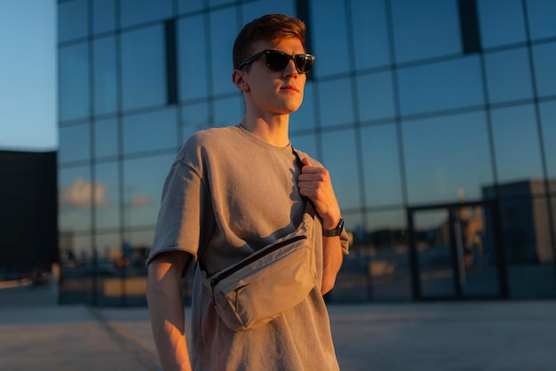 Фото Портрет красивого туриста-хипстера в солнцезащитных очках с сумкой, удобно стоящего парня возле здания в центре города спокойный расслабляющий момент концепции летних каникул