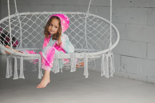 Фото Портрет счастливой девочки на крыльце, сидящей на плетеной качели и играющей в розовой пижаме.