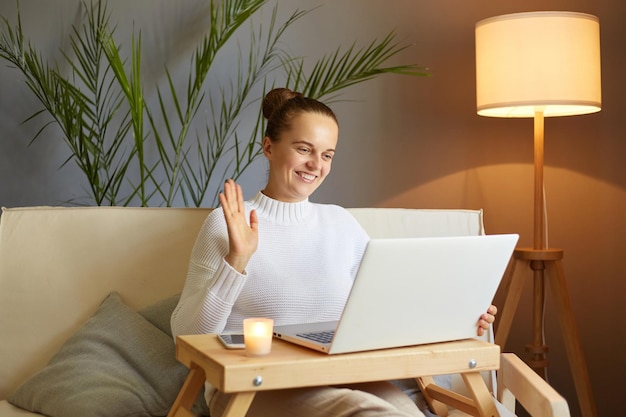 Фото Портрет счастливой довольной кавказской женщины с темными волосами в белом джемпере, сидящей на диване с ноутбуком, проводящей видеозвонок онлайн-встречи, машущей рукой, говоря привет или пока