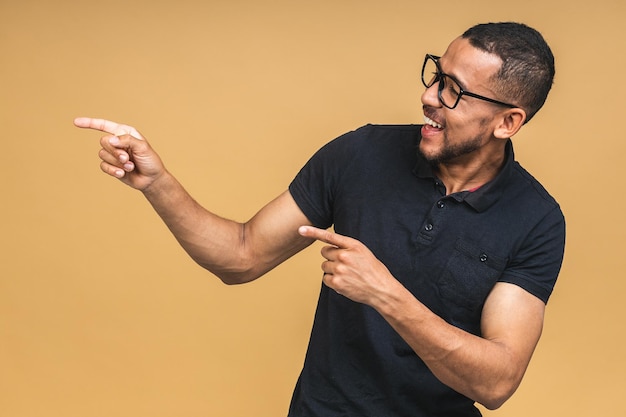 カジュアルな笑みを浮かべてベージュ色の背景に分離された指で脇を指している幸せな若いアフリカ系アメリカ人の黒人男性の肖像画