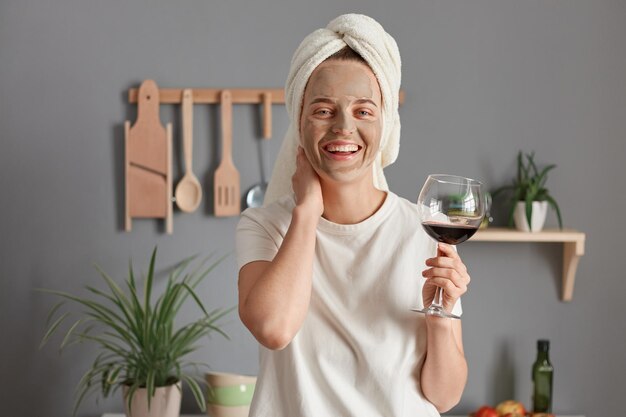 Фото Портрет радостной жизнерадостной женщины с косметической маской в белом полотенце, обернутой вокруг головы, пьющей вино на кухне, держащей руку на шее, смотрящей в камеру с улыбкой, наслаждающейся выходными