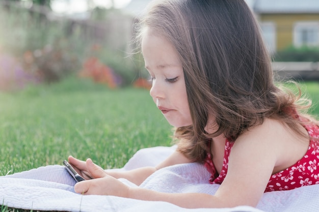 사진 푸른 잔디에 담요에 누워 휴대 전화를 찾고 빨간 옷에 어린 소녀 3-4의 초상화. 가제트를 사용하는 어린이