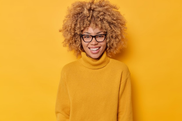 사진 곱슬머리의 사랑스러운 젊은 여성의 초상화는 하얀 치아가 안경을 쓰고 노란색 배경에 격리된 카메라를 직접 바라보는 모습을 행복하게 보여줍니다. 긍정적인 감정 개념입니다.