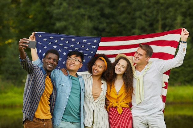 写真 アメリカの国旗を保持し、夏のパーティーを楽しみながら屋外で自分撮りをする友人の多民族グループの肖像画