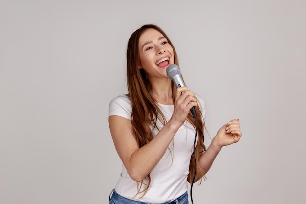 Фото Портрет позитивной привлекательной женщины, громко поющей песню с микрофоном в руке, весело отдыхающей в караоке в белой футболке. внутренняя студия, снятая на сером фоне