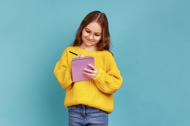 Фото Портрет позитивной маленькой девочки, пишущей в блокноте, улыбающейся заметки школьника, делающего домашнее задание в желтом повседневном свитере. съемка в студии на синем фоне