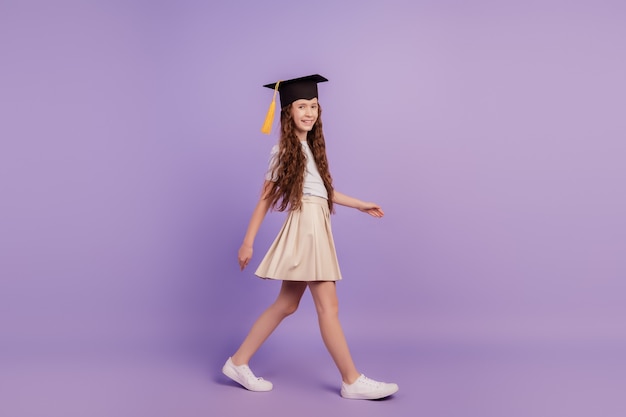 보라색 배경에 졸업 모자를 쓰고 걷는 꽤 작은 학생 소녀의 초상화