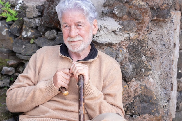 Фото Портрет расслабленного бородатого пожилого мужчины, сидящего на улице дома с тростью в свитере улыбающийся пожилой мужчина смотрит в сторону