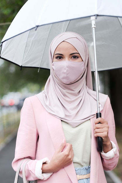 ピンクのヒジャーブと街の通りで傘を保持しているジャケットの宗教的な若い女性の肖像画