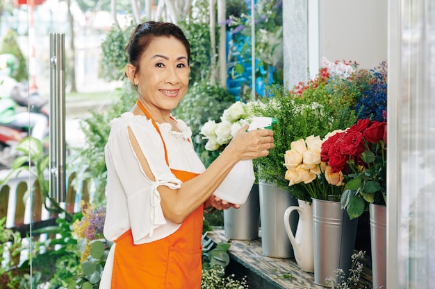 店内の花に水をまくオレンジ色のエプロンでシニアベトナムの花屋の肖像画