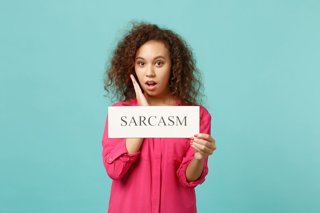 스튜디오의 파란색 청록색 벽 배경에 격리된 Sarcasm 텍스트 보드를 들고 분홍색 캐주얼 옷을 입은 충격을 받은 아프리카 소녀의 초상화. 사람들은 진심 어린 감정, 라이프 스타일 개념입니다. 복사 공간을 비웃습니다.