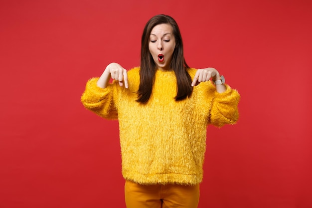 Фото Портрет потрясенной молодой женщины в желтом меховом свитере, указывая указательными пальцами вниз, изолирован на ярко-красном стенном фоне в студии. люди искренние эмоции, концепция образа жизни. копируйте пространство для копирования.