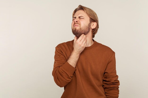 Фото Портрет больного бородатого мужчины, касающегося воспаленных миндалин, сжимающего шею и хмурящегося от боли, страдающего от боли в горле