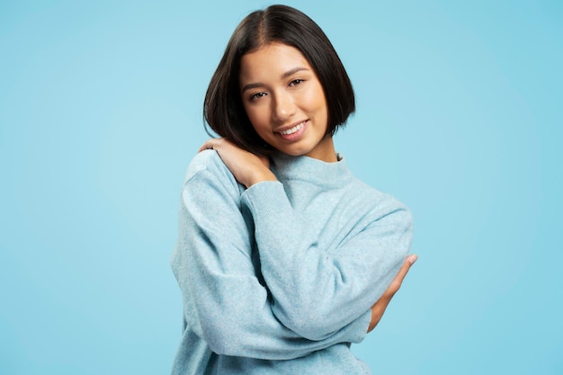 사진 캐주얼 따뜻한 스웨터를 입고 미소 짓는 귀여운 아시아 여성의 초상화. 파란색 배경에 고립되어 서있는 카메라를 바라보며 자신을 포옹하는 쇼핑 개념
