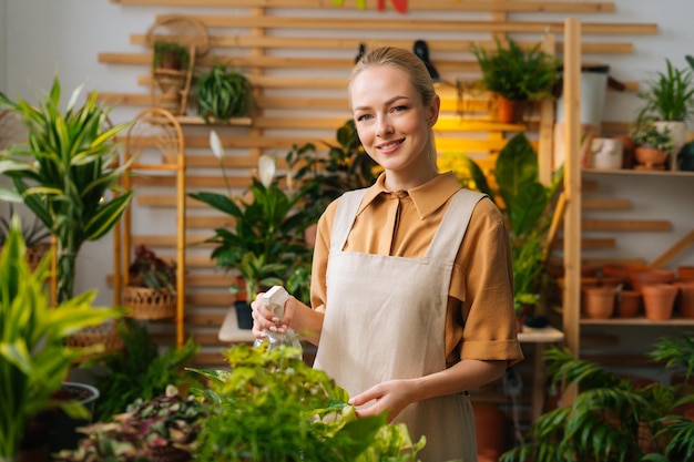 カメラを見ている噴霧器によって植木鉢の観葉植物に水を噴霧するエプロンで笑顔の女性の花屋の肖像画