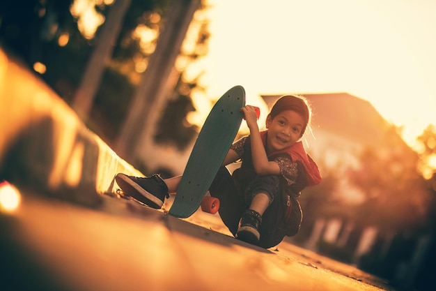 Фото Портрет улыбающейся девушки, держащей скейтборд, сидящей на дороге против неба во время захода солнца