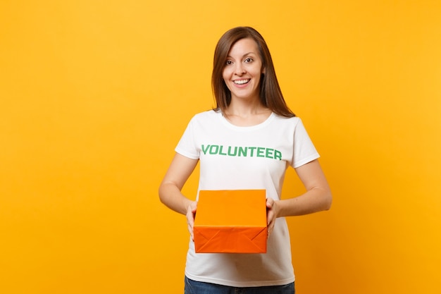 Фото Портрет улыбающейся женщины в белой футболке с надписью зеленый заголовок волонтер держит оранжевую картонную коробку, изолированную на желтом фоне. добровольная бесплатная помощь, концепция благотворительности