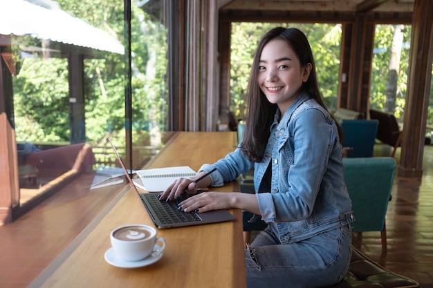 写真 笑顔の若いアジアの女性の肖像画は、コーヒーショップでラップトップで働いていますオンラインで働いています