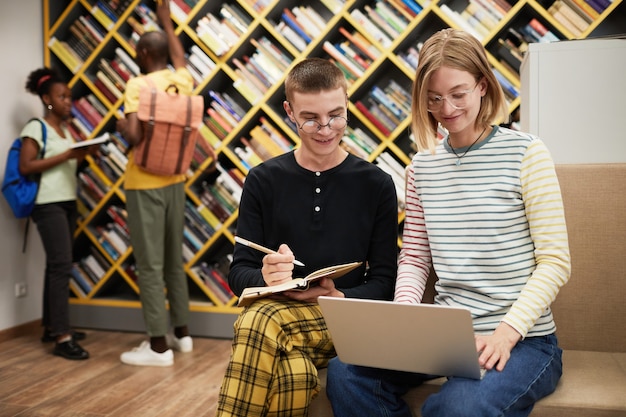 Фото Портрет двух веселых студентов, использующих ноутбук вместе, работая в копировальном пространстве библиотеки колледжа