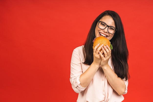 Фото Портрет молодой красивой голодной женщины, поедающей гамбургер изолированный портрет студентки с фаст-фудом на красном фоне концепция диеты
