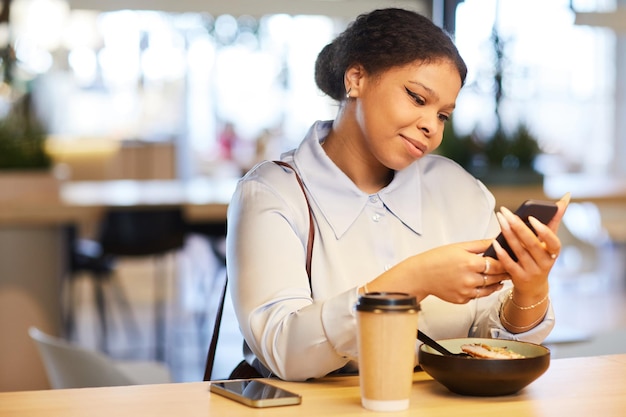 Портрет молодой чернокожей женщины в кафе, смотрящей на экран смартфона и улыбающейся, освещенной теплым светом полицейского