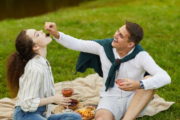 湖のほとりの緑の芝生に座って果物を食べながら屋外でロマンチックなデートを楽しんでいる若いのんきなカップルの肖像画