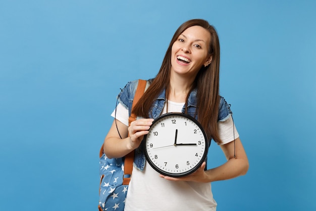若いカジュアルな笑い楽しい女性学生の肖像画tシャツ、青い背景で隔離の目覚まし時計を保持しているバックパックとデニムの服。大学での教育。広告用のスペースをコピーします。