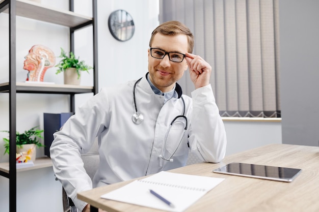 Фото Портрет молодого врача в очках и белой форме со стетоскопом, говорящего и консультирующего пациента онлайн, смотрящего в камеру, делающего видеозвонок, сидя за столом в офисе