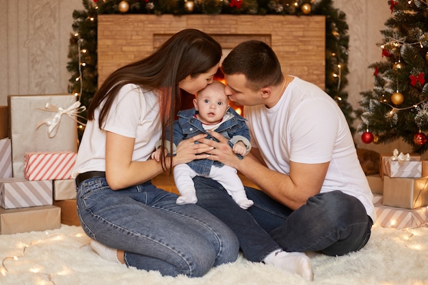 幼い娘にキスをし、クリスマスの木と暖炉の近くの床に座って、家で一緒に冬の休暇を過ごし、祝う若い家族の肖像画。