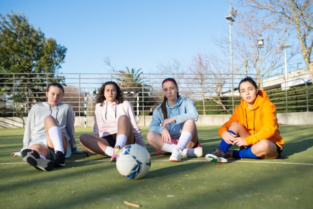 사진 그린 필드에 젊은 여성 축구 선수의 초상화입니다. 운동복을 입은 4명의 진지한 소녀들이 훈련을 하고 카메라를 바라보기 전에 바닥에 앉아 있습니다. 팀 스포츠와 건강한 라이프 스타일 개념