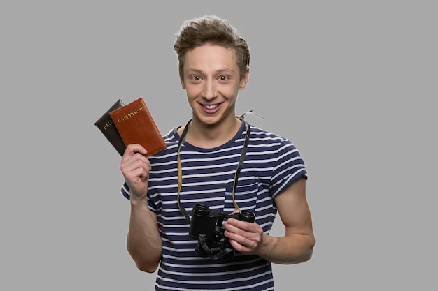 Фото Портрет молодого счастливого мальчика, держащего паспорта и бинокль. туристический парень показывает паспорта на сером фоне. концепция путешествия.