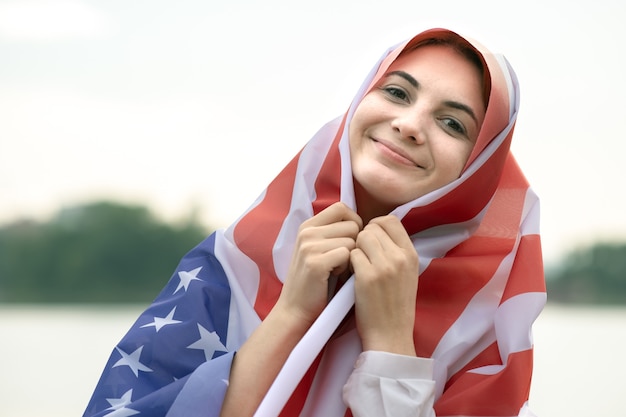 그녀의 머리와 어깨에 미국 국기와 함께 젊은 행복 한 난민 여성의 초상화. 미국 독립 기념일을 축하하는 긍정적인 이슬람 소녀.