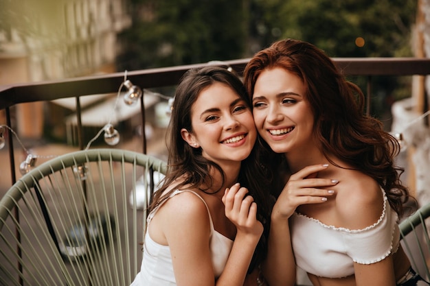 Фото Портрет юных леди в белых нарядах, позирующих на балконе две милые подруги расслабляются и мило улыбаются на террасе в легкой одежде