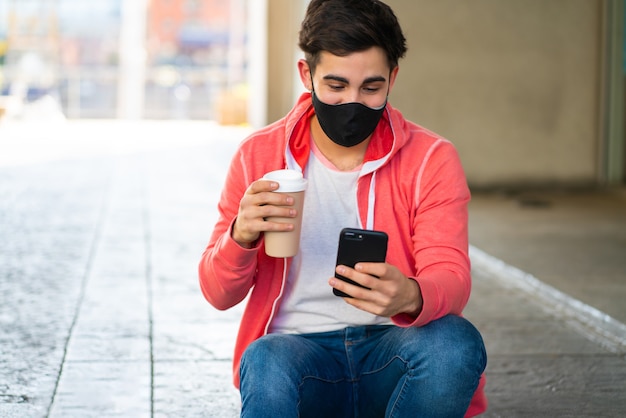 彼の携帯電話を使用して、路上で屋外に座ってコーヒーを飲む若い男の肖像画。フェイスマスクを着用した男。アーバンコンセプト。