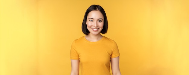 Фото Портрет молодой современной азиатки, счастливо улыбающейся с белыми зубами, уверенно смотрящей в камеру в повседневной футболке, стоящей на желтом фоне