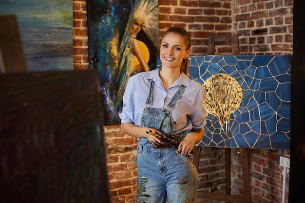 Фото Портрет молодой улыбающейся художницы с ее работами в художественной студии художник держит художественные кисти ...