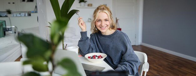 Фото Портрет молодой улыбающейся женщины, сидящей в комнате и смотрящей видео на ноутбуке, едя обед или завтрак