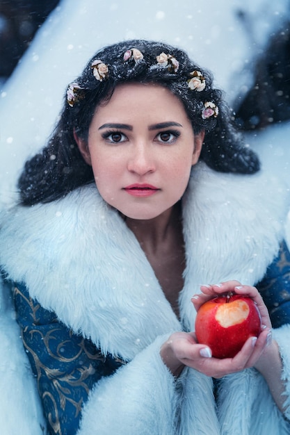 写真 青いコートを着た若い女性の肖像画。彼女はリンゴの入ったバスケットを持って冬の森を歩いています
