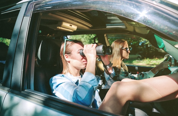 彼女の友人がロードトリップの冒険で運転している間双眼鏡を通して見ている若い女性の肖像画。女性の友情と余暇の概念。